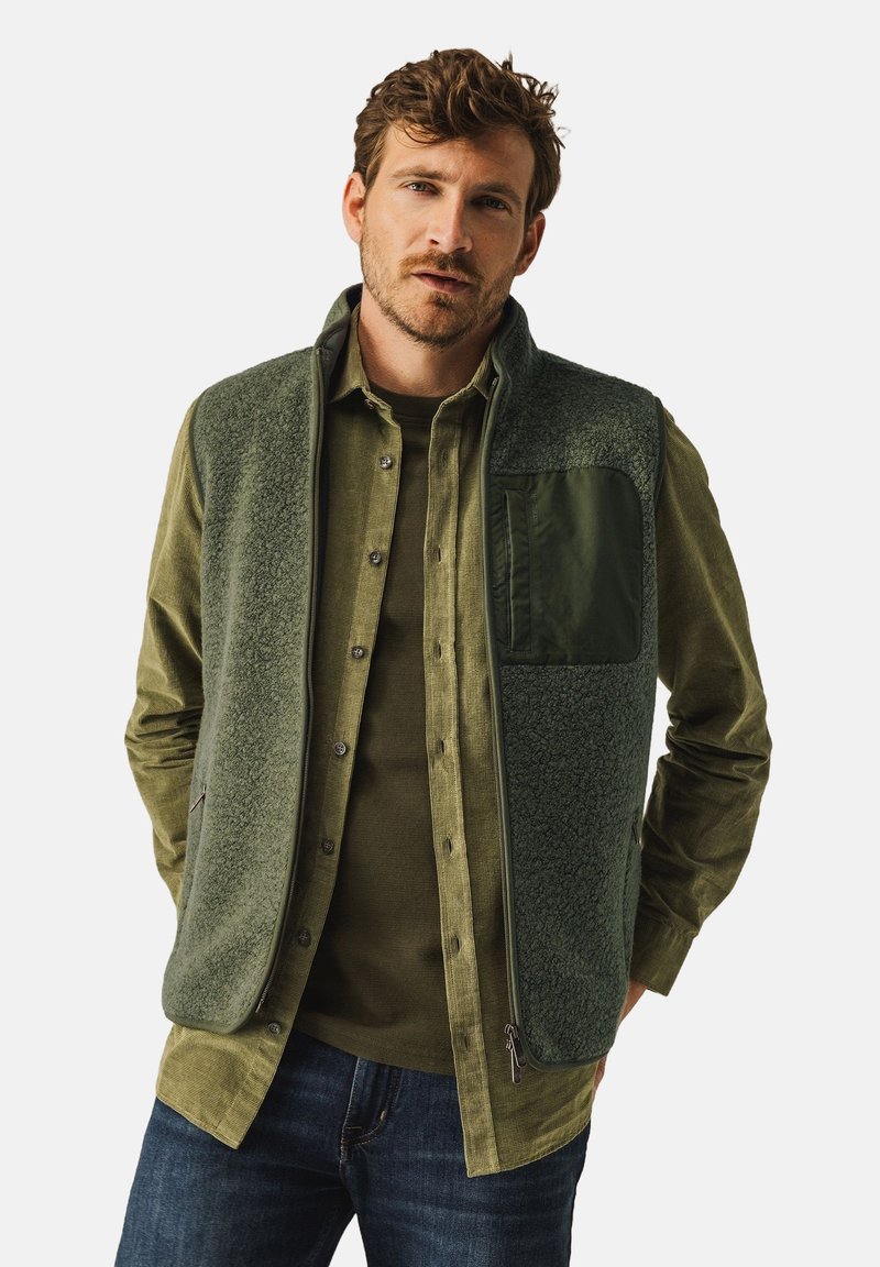 Green fleece vest with a zip closure, textured front, and a solid green pocket. Worn over a green button-up shirt, paired with dark jeans.