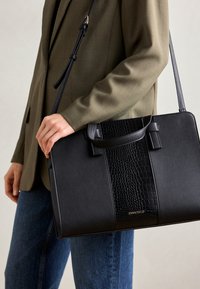 Sac à main noir avec un motif crocodile texturé, des côtés lisses, des poignées courtes et une bandoulière détachable ; il arbore un logo doré.