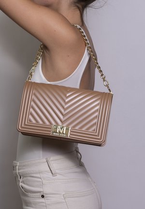 Vrouw in wit topje en lichte broek draagt een beige doorgestikte handtas met gouden kettingriem over haar schouder tegen een grijze achtergrond.