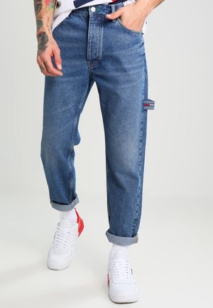 Mężczyzna noszący podwinięte niebieskie jeansy z małą boczną pętlą, białe trampki z czerwonymi akcentami oraz widoczne tatuaże na lewym przedramieniu.