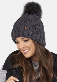 Bonnet en tricot gris avec des câbles texturés, doté d'un grand pompon en fausse fourrure noire sur le dessus et d'une petite étiquette de logo marron sur le côté.
