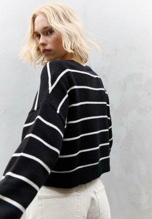Schwarz-weißer gestreifter Strickpullover mit cropped Design und geripptem Saum, kombiniert mit hellen Hosen, vor einem neutralen Hintergrund.