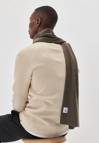 Mand sidder på en træstol iført beige sweater, mørke bukser og et brunt tørklæde draperet over skuldrene, med ryggen til på ensartet baggrund.