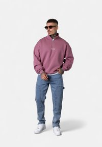 Sweatshirt rose à col zippé avec texte brodé, associé à un jean cargo bleu clair avec poches et des baskets blanches. Les accessoires comprennent des lunettes de soleil.