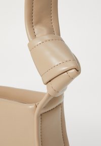 Beżowy skórzany uchwyt torebki z zaokrąglonym wierzchem, z widocznymi przeszyciami i węzłem, który dodaje tekstury i interesującego designu.