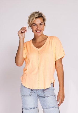 Smiling woman with short blonde hair wearing a peach V-neck shirt and light blue denim shorts, raising her right hand near her face.