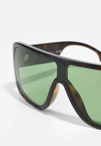 Occhiali da sole tinti di verde con una montatura spessa, angolare, nera e a tartaruga. Superficie liscia e leggermente lucida con un design avvolgente.