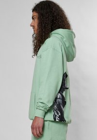 Šviesiai žalia džemperis su gobtuvu, ant šono turintis didelį juodą grafikos spaudą. Ilgos rankovės, priekinė kišenė ir minkštas audinio tekstūros jausmas.