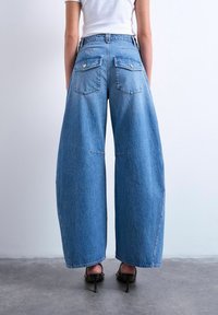 Femme portant un jean bleu à jambes larges et taille haute avec poches arrière, un haut blanc ajusté et des sandales à talons hauts noires, debout sur un sol gris.
