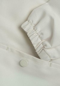 Tissu beige clair avec une texture subtile. Présente une manche froncée et un bouton blanc avec "Chicco" embossé dessus.