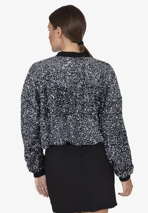 Donna con i capelli castani legati all'indietro, che indossa una giacca nera con paillettes e una gonna nera, rivolta di spalle rispetto alla telecamera su uno sfondo bianco.