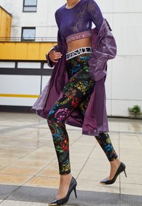 Top lungo a maniche trasparenti viola, leggings neri con motivo paisley multicolore e trench coat lucido viola, abbinati a tacchi neri.