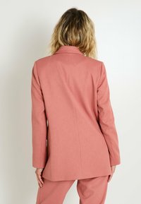 Blazer ajusté rose pâle avec un col cranté, des manches longues et des détails de couture simple, associé à un pantalon assorti.