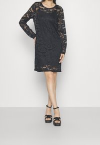 Černé krajkové šaty s dlouhými rukávy a kulatým výstřihem, zdobené květinovým vzorem a mírně průsvitnou texturou. V kombinaci s černými platformovými sandály.