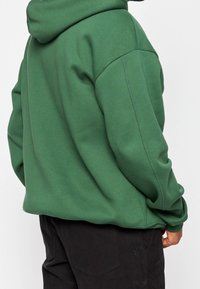 Felpa verde realizzata in tessuto morbido, con cappuccio, spalle cadute e polsini a costine. Abbinata a pantaloni neri, con dettagli di cucitura minimali.