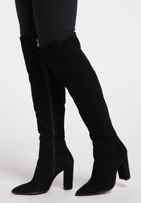 Stivali in suede neri sopra il ginocchio con punta affilata, tacco a blocco e dettagli con zip sul lato, che mostrano una texture liscia e una silhouette elegante.