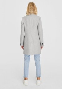 Cappotto di lana grigio con taglio dritto, apertura singola sul retro e dettagli con bottoni neri, abbinato a jeans azzurri e sneakers bianche.