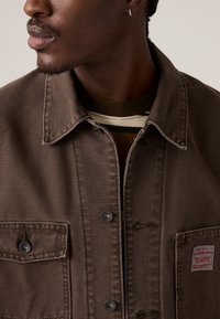 Hombre que lleva una chaqueta de mezclilla marrón con botones de metal y una camiseta a rayas debajo, visto de los hombros hacia arriba.
