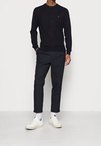 Schwarzer Strickpullover mit Rundhalsausschnitt, kombiniert mit dunkler Hose. Weiße Sneaker mit beiger Sohle. Einfaches, lässiges Design, ohne Muster oder Akzente.