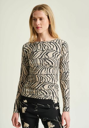 Mujer con cabello rubio largo que lleva una camiseta de manga larga de estampado de cebra en beige y negro y pantalones negros con patrones abstractos claros, de pie mirando ligeramente hacia la izquierda.