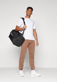 Sort duffel taske med mesh-detaljer, båret over skulderen; hvid crew neck t-shirt; brune tapered joggers; hvide sneakers.