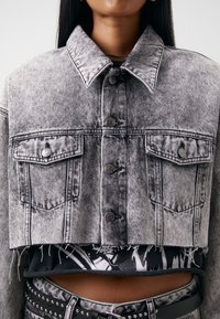 Lyhyt harmaa farkkutakki, jossa on kaksi etupokketa, napitus ja revitty helma, kerroksittain mustan graafisen paidan päällä.