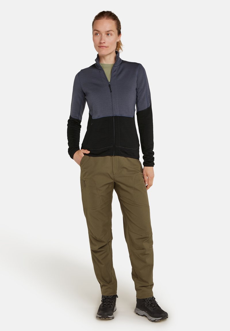 Frau in einer dunkelblauen und schwarzen Zip-Jacke, olivegrünen Hosen und schwarzen Wanderschuhen, stehend mit einer Hand in der Tasche vor weißem Hintergrund.