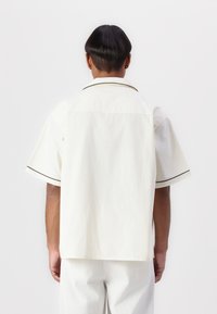 White short-sleeve shirt with a relaxed fit, featuring a black piped collar and contrasting cuffs. Smooth cotton fabric with a subtle texture.
