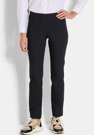 Pantalon noir ajusté avec une texture lisse, taille haute et poches latérales, associé à des sneakers beiges et noires.
