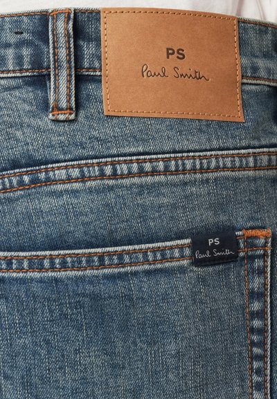 Mėlynos džinsinės kelnės su ruduo odiniu ženklu, pažymėtu "PS Paul Smith." Savybė - oranžinės siūlės ir maža tamsiai mėlyna etiketė ant galinio kišenės.