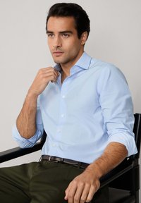 Camisa de botones azul claro con mangas largas y un patrón texturizado, combinada con un cinturón oscuro y pantalones verde olivo, sentado contra un fondo neutral.