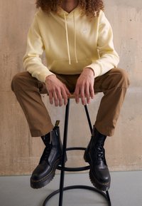 Gul huva tröja med framficka och vitt logotyp, matchad med bruna byxor och svarta snörskor med tjock sula.