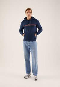 Felpa navy con tasca frontale, dettaglio a righe in rosso e verde, abbinata a jeans azzurri chiari e sneakers argentati. Texture del tessuto liscia.