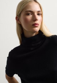 Maglione nero a collo alto con texture morbida, maniche corte e una vestibilità aderente sul colletto, presentato su uno sfondo neutro.