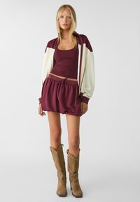 Maroon atletisk crop top och matchande lösa shorts, parat med tan cowboy stövlar. Jackan är krämfärgad med maroon accenter och en sportig design.