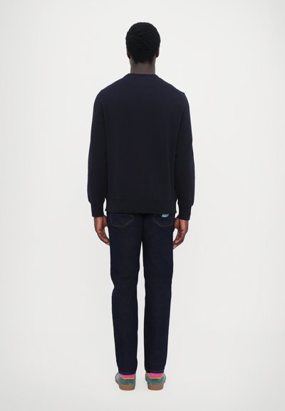 Sweatshirt marine avec des poignets et un ourlet côtelés, porté avec un jean en denim foncé. Les chaussures présentent des accents multicolores et une semelle texturée.