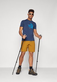 Navy t-shirt with blue graphic design, paired with mustard shorts. Accessories include black hiking poles and rugged black hiking boots.