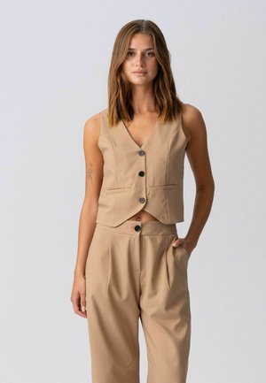 Vállig érő barna hajú nő, bézs színű, ujjatlan, gombos mellényt és hozzáillő, magas derekú nadrágot visel, egyik kezét zsebre dugva.