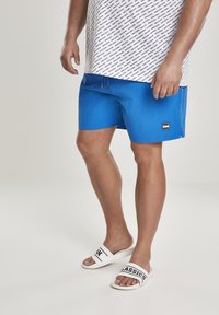 Shorts de bain bleus avec une texture lisse, taille à cordon de serrage et un patch logo. Associés à des claquettes blanches avec une bande imprimée noire.