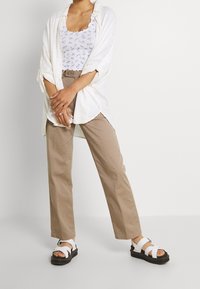 Vit skjorta med knappar ovanpå en blommig linne, ihop med beige raka byxor och vita sandaler med svarta sulor.