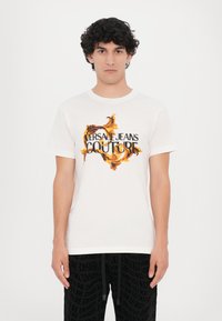 Balta medvilninė t-shirt, puošta ryškiomis gėlių grafikomis ir juodu tekstu "VERSACE JEANS COUTURE". Trumpomis rankovėmis, klasikinis apvalus kaklas.