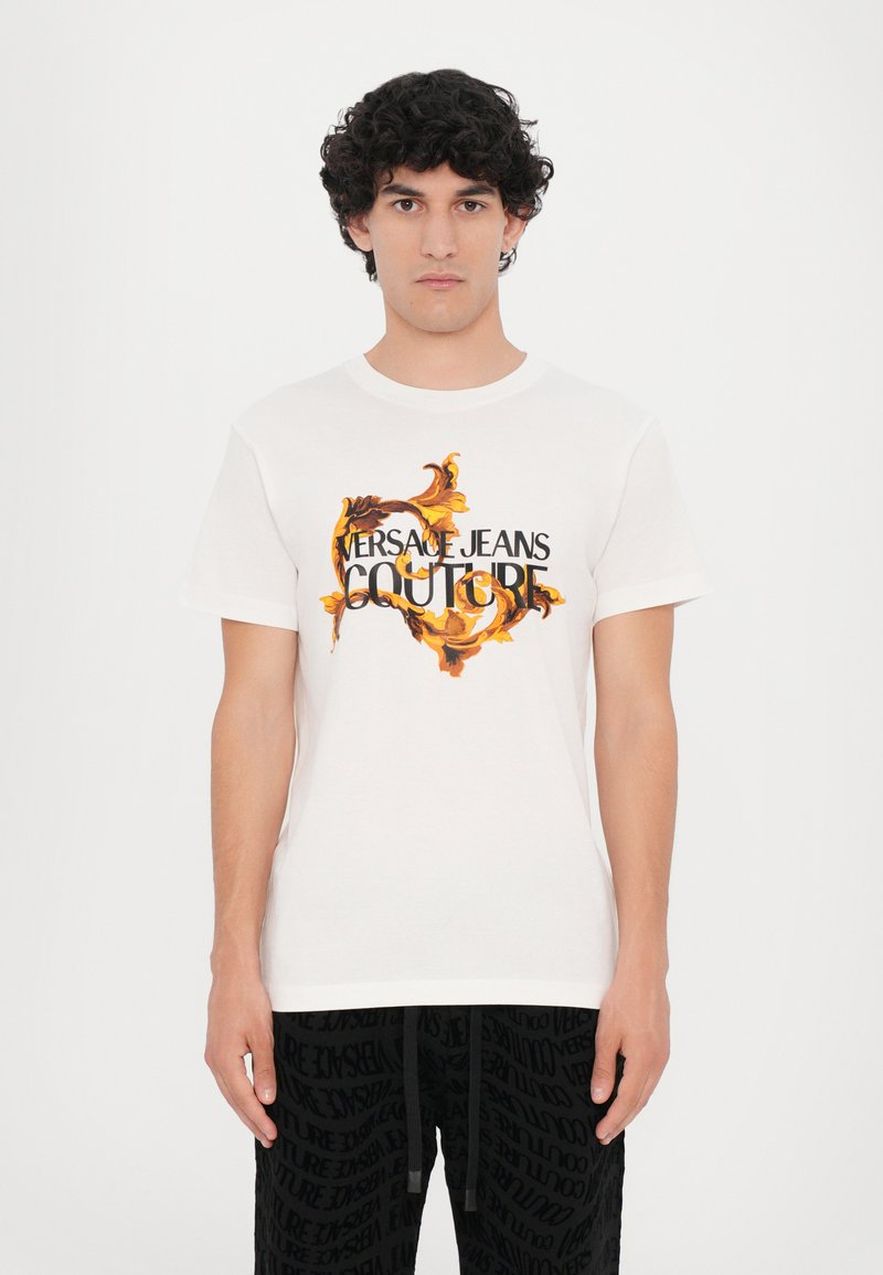 Balta medvilninė t-shirt, puošta ryškiomis gėlių grafikomis ir juodu tekstu "VERSACE JEANS COUTURE". Trumpomis rankovėmis, klasikinis apvalus kaklas.