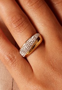 Bague en or avec un anneau lisse et une bande diagonale de petites cristaux transparents. Peau texturée visible en arrière-plan.