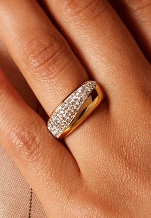 Gouden ring met een gladde band en een diagonale strook kleine heldere kristallen. Textuur van huid zichtbaar op de achtergrond.