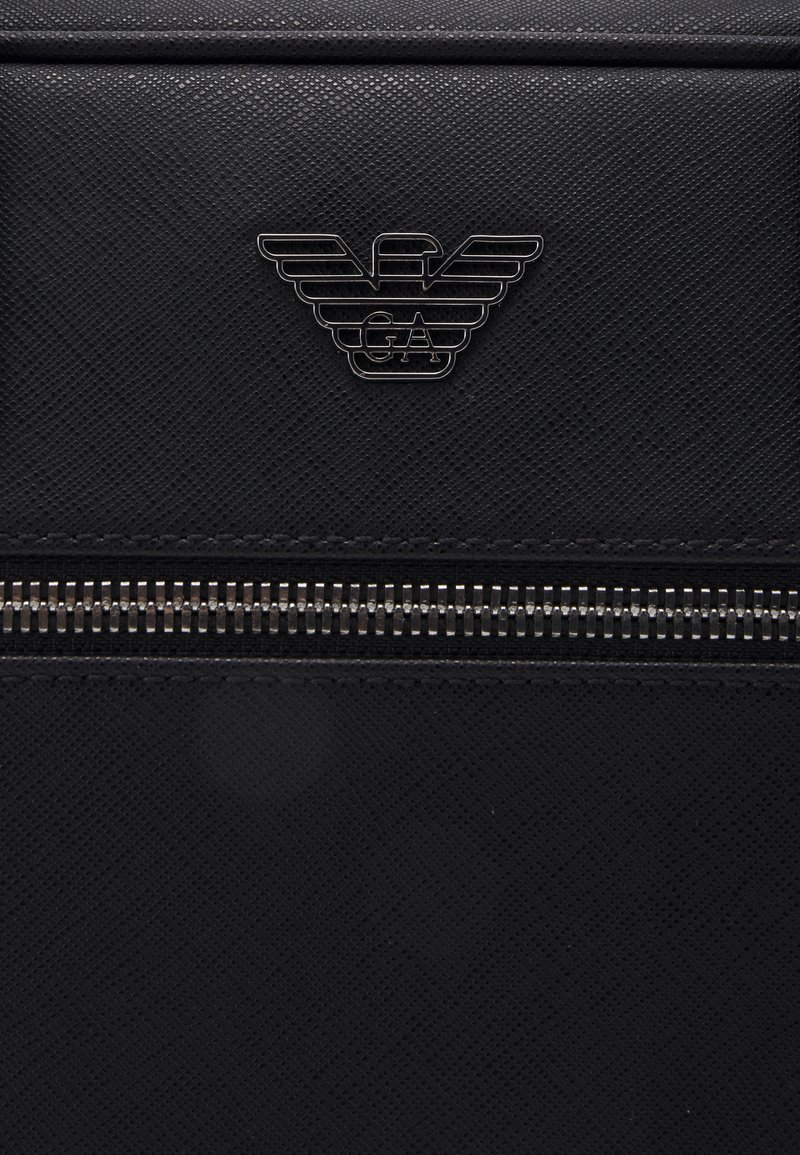 Sac en cuir texturé noir avec une fermeture éclair argentée, arborant un logo en métal noir en haut. Le design est épuré et minimaliste.