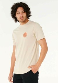 Beige T-shirt med korte ermer og et rundt orange logo på venstre bryst. Blødt stof, tætsiddende design, rund hals og lige kant.