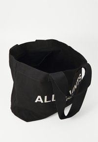 Borsa tote nera in tela con apertura ampia, dotata di spallacci robusti e testo bianco audace che recita "ALL SAINTS" su un lato.