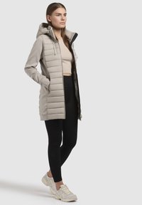 Beige isoleret jakke med lynlås foran og hætte, med glat stof på ærmerne, og parret med sorte leggings og lysfarvede sneakers.