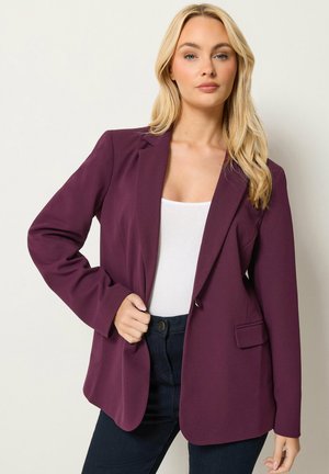 Femme blonde portant un blazer violet par-dessus un top blanc et un jean foncé, debout avec une main tenant le revers du blazer.