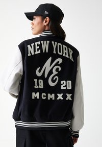 Black and white varsity jacket featuring "NEW YORK" and "MCMXX" in felt embroidery, with ribbed cuffs and a white sleeve.